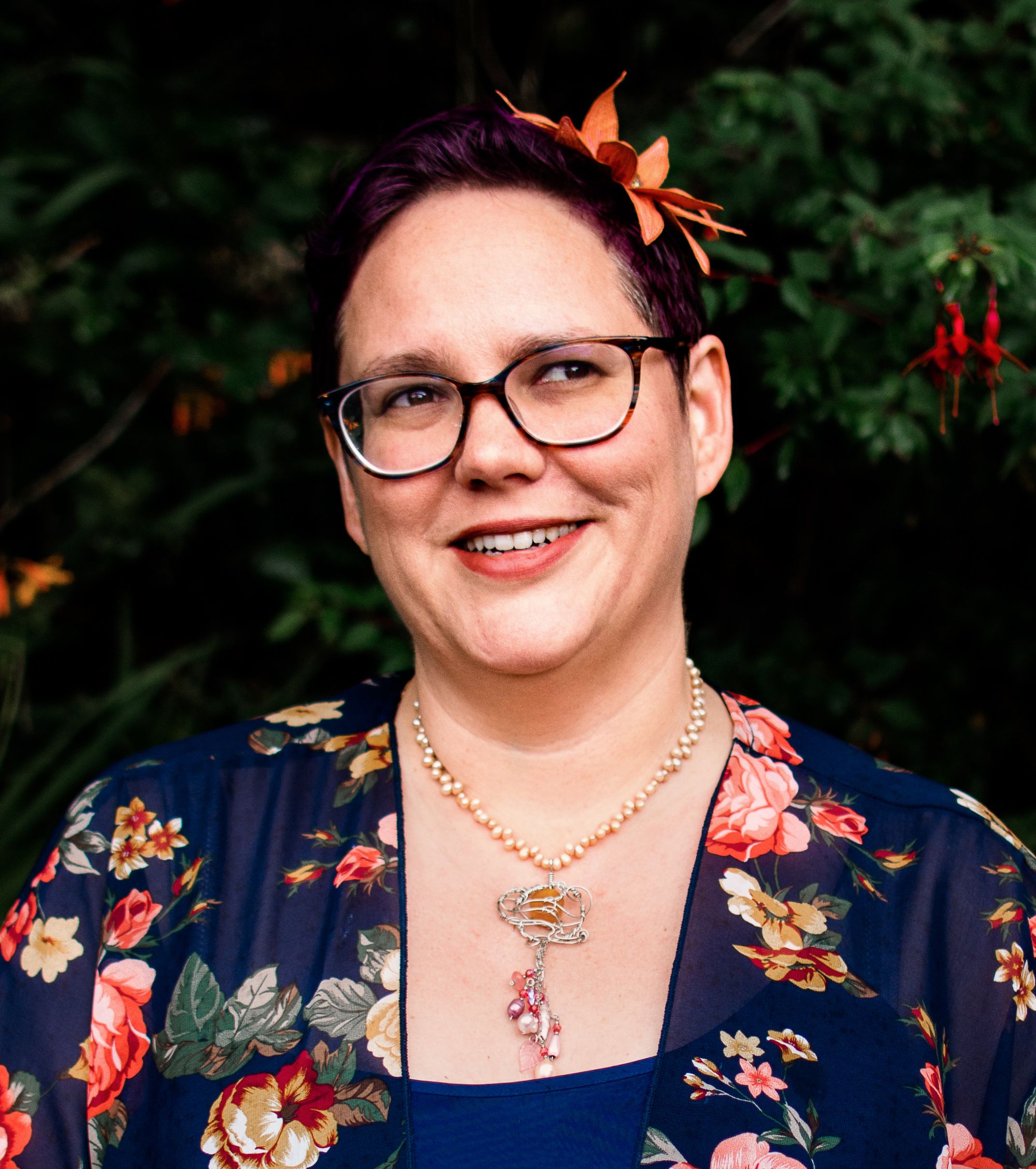 Me! A person with short purple hair and an orange hair flower (made by Betsie Withey), wearing a navy blue floral top and glancing upward as if they have a mischievous idea, perhaps? Photo by Marie O’Mahony Photography, August, 2019.
