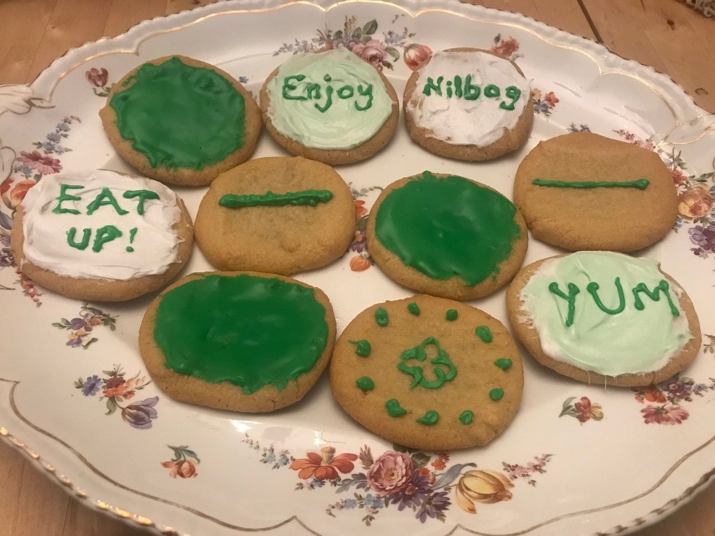 a floral ceramic platter full of cookies decorated with green icing. Some are plain, some have one green line, one has a clover, and four have words: Yum, Enjoy, Nilbog, and Eat Up!