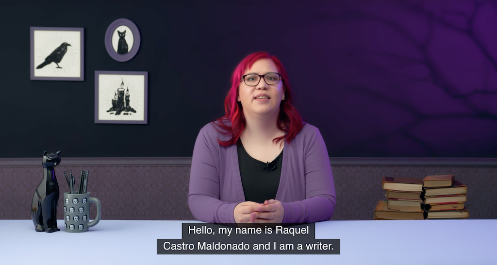 Raquel Castro, a woman with long fuchsia hair, sits in a a room with purple walls and horror themed decor including black cats, ravens, and an Edgar Allen Poe mug full of pens. The subtitle says, "Hello, my name is Raquel Castro Maldonado and I am a writer."