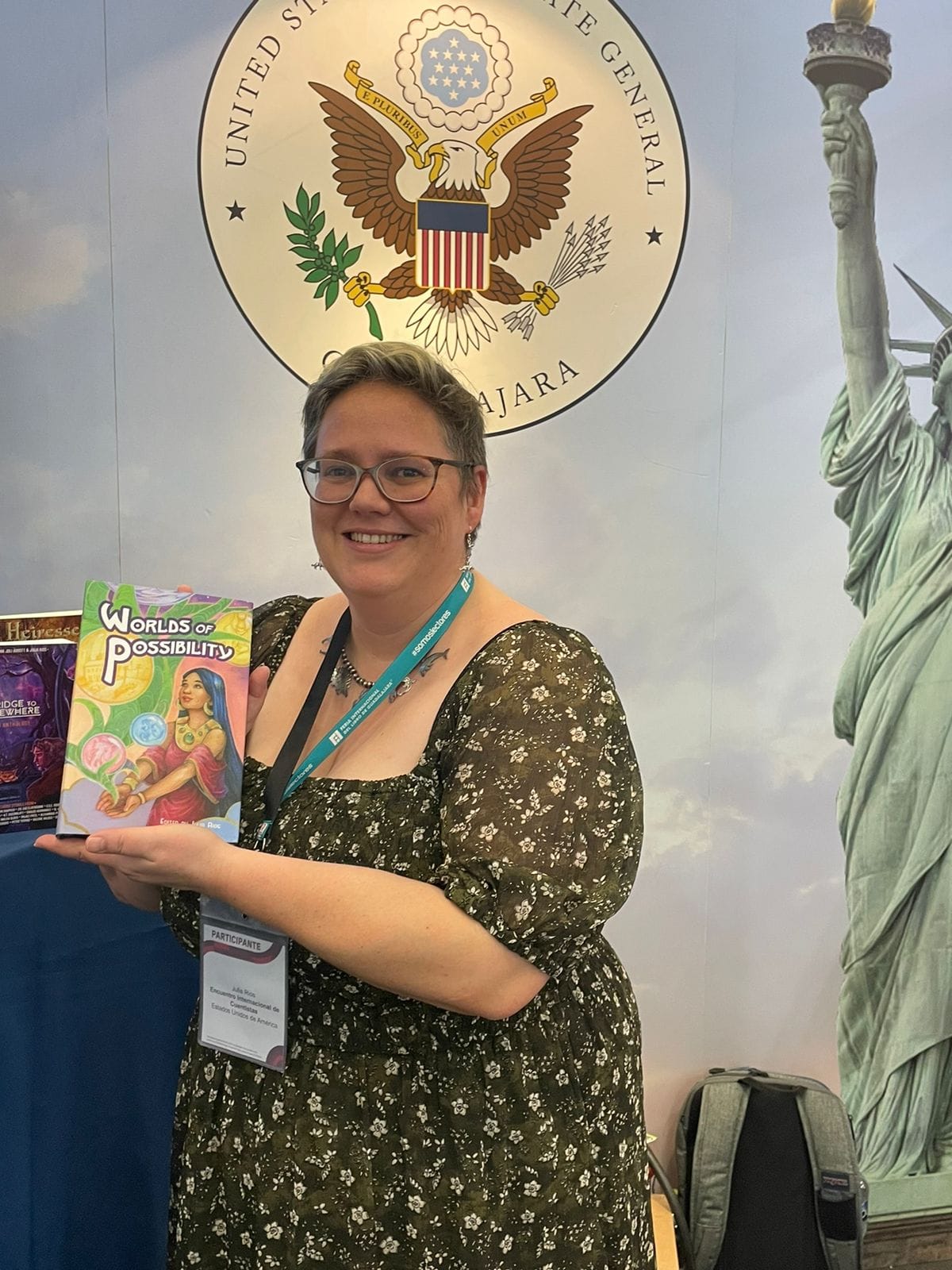 Julia Rios in in a floral dress, holding a copy of the hardcover edition of Worlds of Possibility. They are stanging in front of a backdrop with the seal of the United states and the Statue of Liberty.