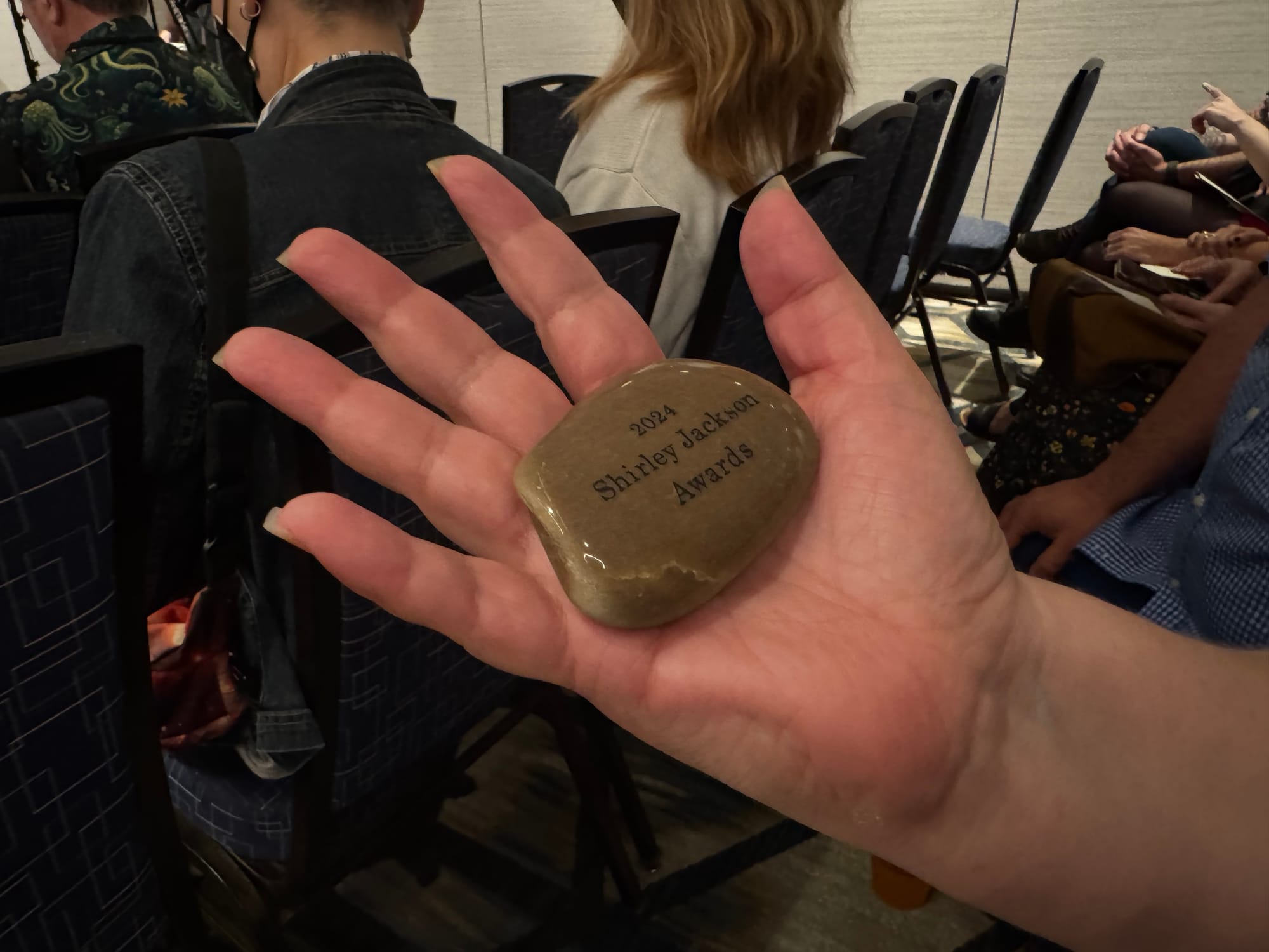 a polished rock engraved with "2024 Shirley Jackson Awards" rests in an open hand.
